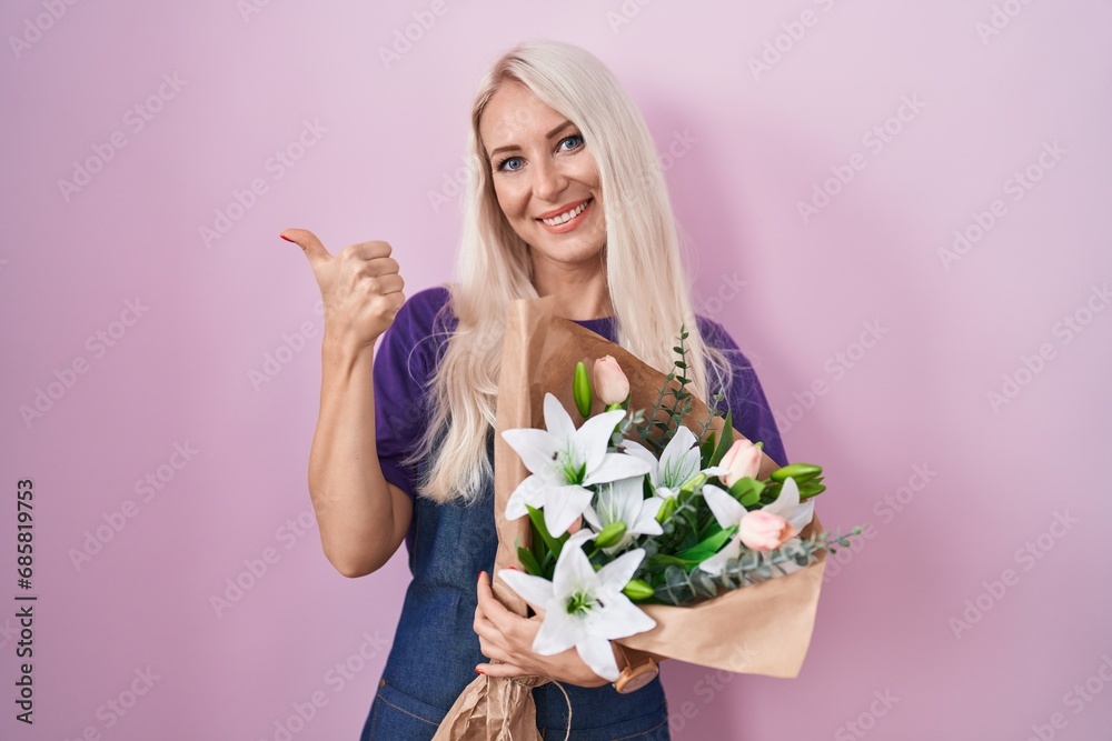 Caucasian woman holding bouquet of white flowers pointing to the back behind with hand and thumbs up, smiling confident