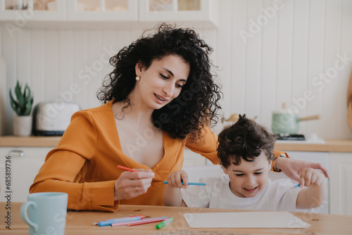 Creative little curly boy drawing at home sitting with mom at desk at kitchen. Family domestic leisure, childhood. Cheerful young hispanic woman spending time with son. Babysitter with kid.