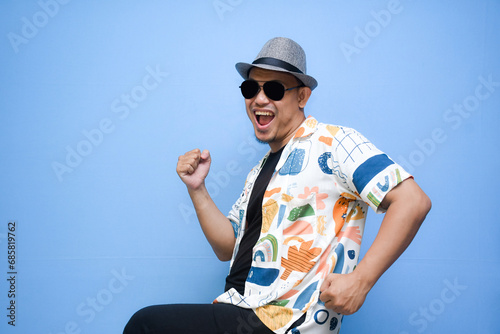 Overjoyed asian man wearing sunglasses dancing, smiles positively, being in high spirit. isolated on blue background. holiday and vacation concept