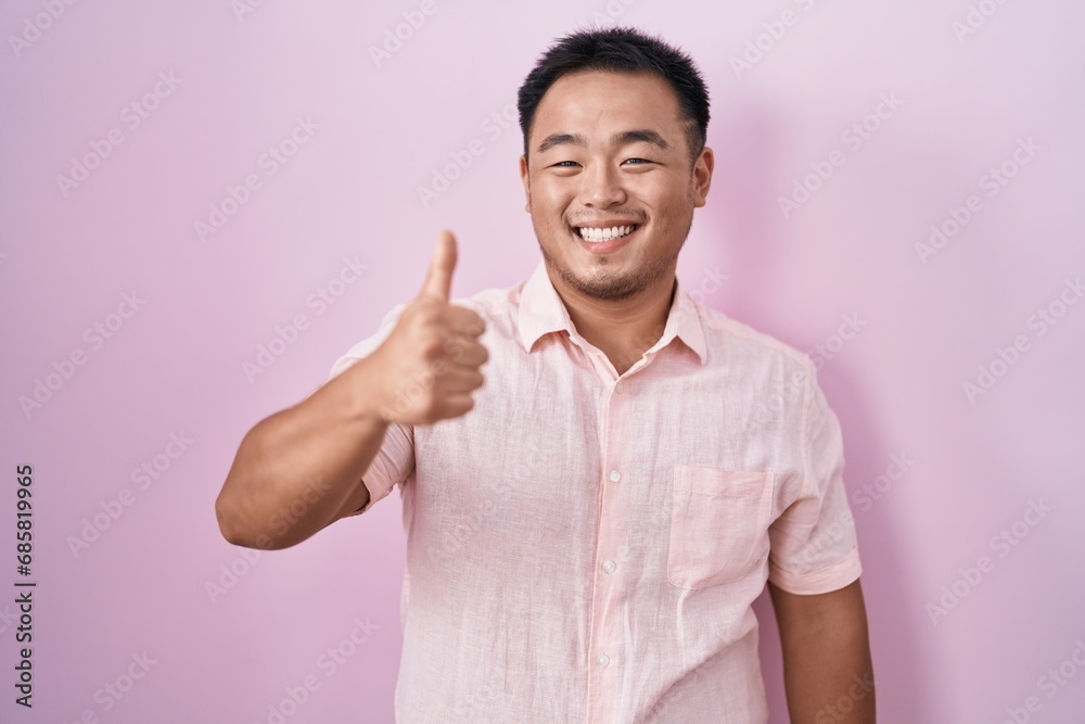 Chinese young man standing over pink background doing happy thumbs up gesture with hand. approving expression looking at the camera showing success. Chinese young man standing over pink background doing happy thumbs up gesture with hand. approving expression looking at the camera showing success.