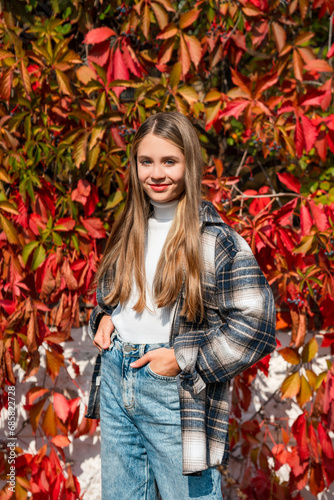 Portrait of a beautiful girl in a shirt walking in the park against the background of a bush with red autumn leaves, a cozy autumn atmosphere