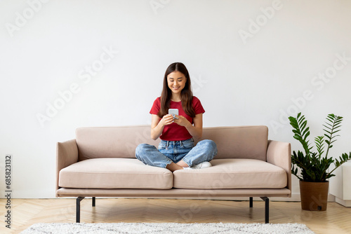 Smiling Young Asian Woman With Smartphone Relaxing On Couch At Home © Prostock-studio