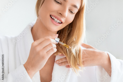 Blonde lady in bathrobe applies nourishing serum to hair indoor