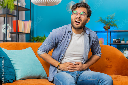 Indian Arabian man sits on sofa feeling sudden strong abdominal stomach ache gastritis problem. Bearded Arabian guy having symptom poisoning diarrhea indigestion peptic ulcer pancreatitis at home room photo