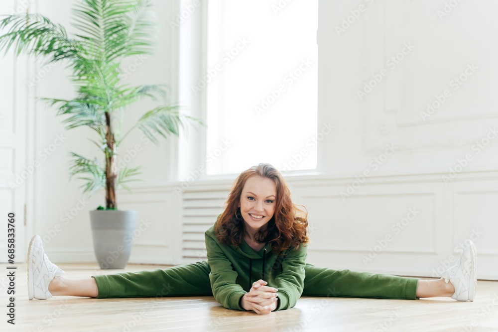 Joyful woman in green sportswear performing splits on white floor, fitness at home, bright interior with potted plant