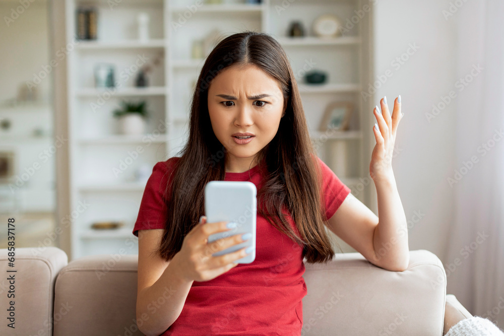 Displeased young asian woman looking at smartphone screen at home Displeased young asian woman looking at smartphone screen at home
