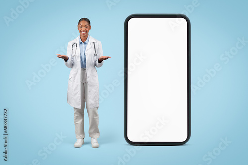 Cheerful black young woman doctor in white coat, hold free space on hands near big phone