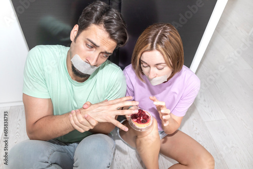 Hungry couple with taped mouths sitting close to the refrigerator and trying to keep their diet safe 