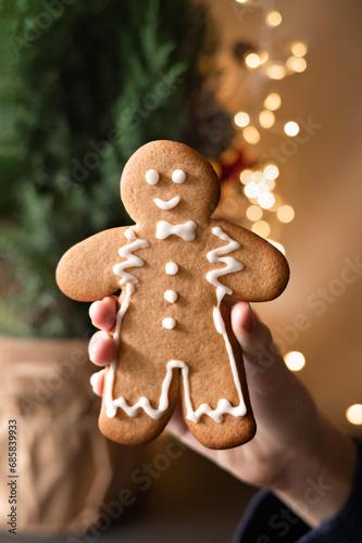 Big gingerbread man cookie in hand, blurred background with Christmas tree in pot and garland lights. Family winter holiday celebration, traditional Christmas treat for child. Soft selective focus