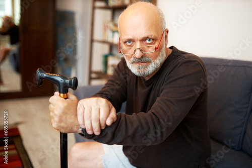 Indoor portrait of angry senior bearded bald man in shorts and long-sleeve sitting on couch looking at camera over eyeglasses, holding walking stick, frustrated with arthritis and staying at home