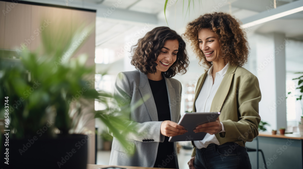 Two professional women smiling and looking at a tablet together in an office setting Two professional women smiling and looking at a tablet together in an office setting