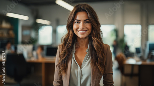 smiling white woman in tan blazer stands confidently in office with people and equipment, creating positive, professional atmosphere