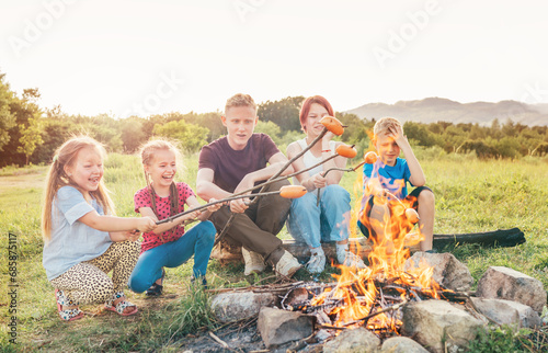 Five kids group Boys and girls cheerfully laughed and roasted sausages on sticks over a campfire flame near the green tent. Outdoor active time spending or camping in Nature concept.