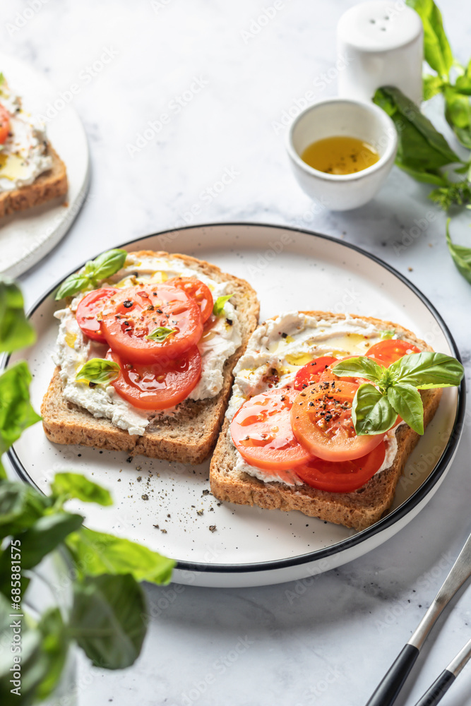 Two sandwiches or toasts with tomatoes, cream cheese, olive oil and basil on a plate on white marble background. Mediterranean breakfast