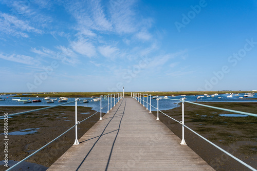 Empty walkway over blue ocean  coastline and horizon seen a large long wooden jetty with a thin white metal railing along the edges but in the perspective distance a blue sky and an ocean with boats.