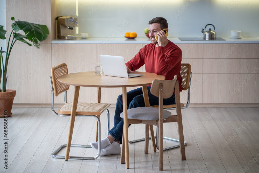 Positive cheerful middle-aged man working at laptop computer in cozy apartment, speaking on mobile phone during work break, solving business issues, sharing ideas on job projects with colleagues