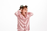 Cute little girl with curlers, in pink silk pajamas on a white background holds her hands behind her head and laughs