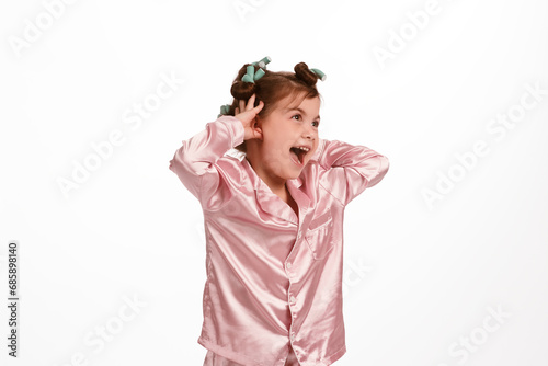 Cute little girl with curlers, in pink silk pajamas on a white background holds her hands behind her head and laughs