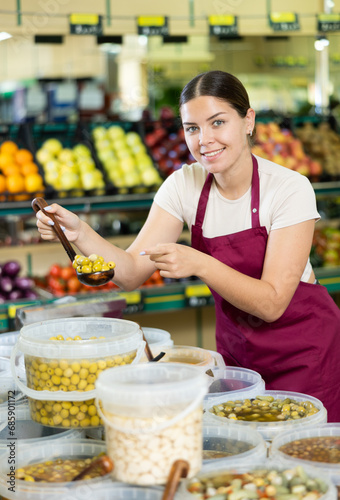 Young woman seller in apron lays out olives in supermarket