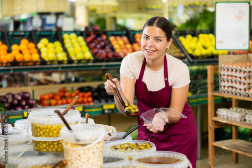 Positive female supermarket grocery clerk laying pickled olives in a container
