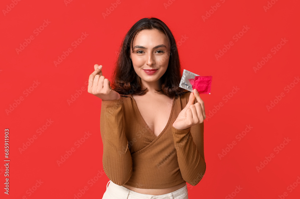 Young woman making heart shape with her fingers and holding condoms on red background. Safe sex concept Young woman making heart shape with her fingers and holding condoms on red background. Safe sex concept