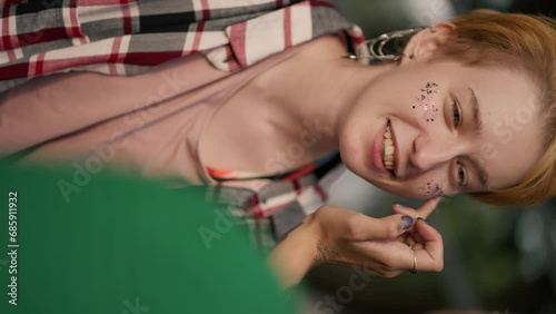 Over the shoulder: vertical video shooting close up, a girl in a green T-shirt applies glitter to her girlfriend with a blonde with a short haircut in a checkered shirt using her fingertip in the park