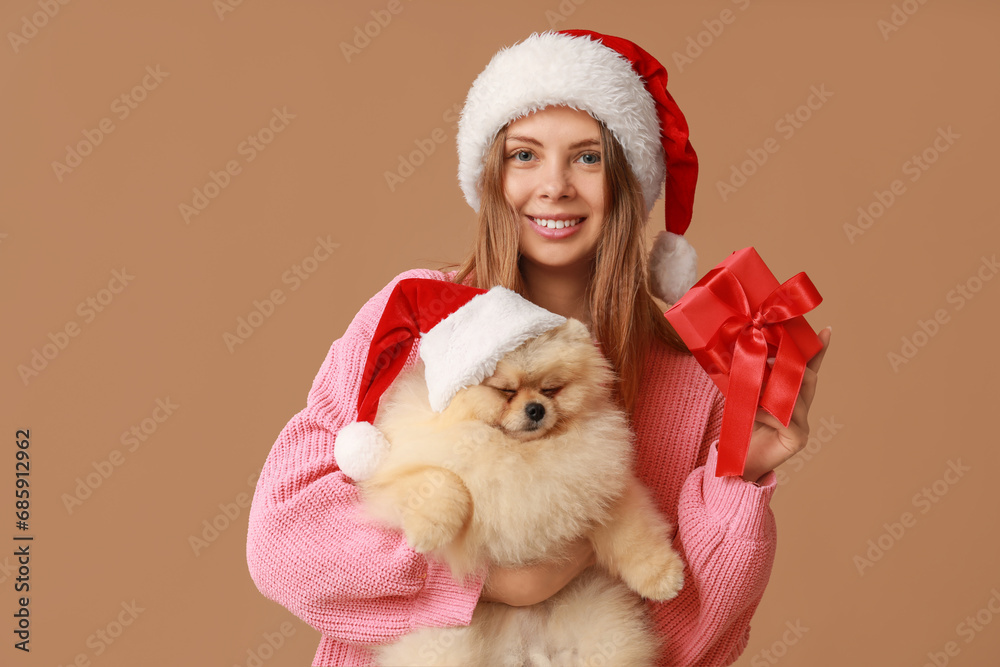 Young woman with cute Pomeranian dog in Santa hats and Christmas gift box on brown background Young woman with cute Pomeranian dog in Santa hats and Christmas gift box on brown background