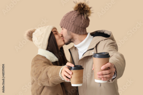 Loving couple in winter clothes with coffee cups kissing on beige background