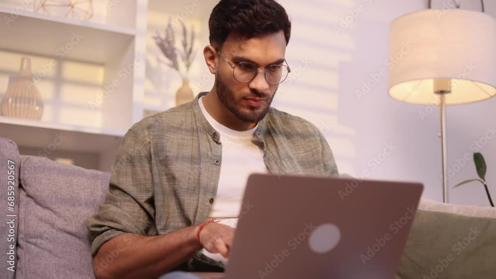 Handsome man with glasses browse products in internet online shopping store planning trip looking for job or swiping through profiles on dating service on laptop computer sitting on couch at home Handsome man with glasses browse products in internet online shopping store planning trip looking for job or swiping through profiles on dating service on laptop computer sitting on couch at home