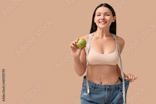 Beautiful young happy woman in loose jeans with fresh apple and measuring tape on beige background. Weight loss concept