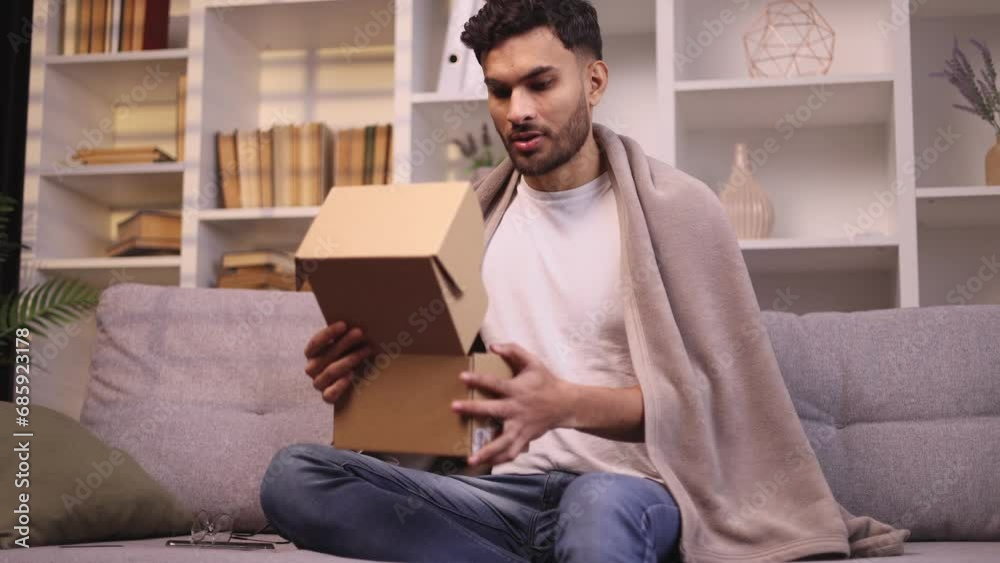 Handsome young bearded man received parcel satisfied with shopping in online internet store looking into box at home Happy customer rejoice with purchase order indoors Handsome young bearded man received parcel satisfied with shopping in online internet store looking into box at home Happy customer rejoice with purchase order indoors