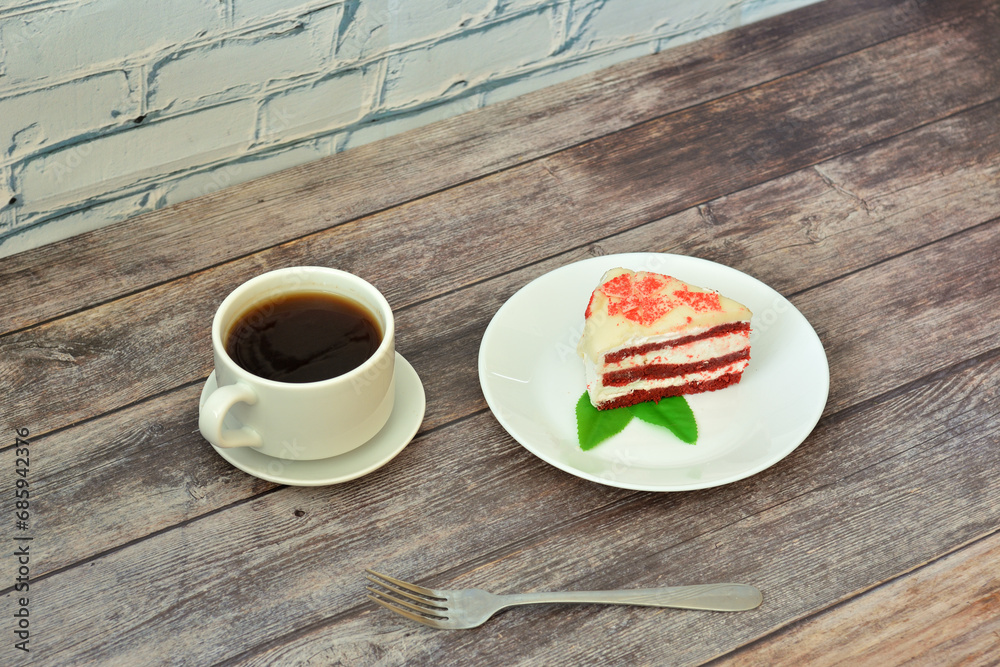 Obraz premium A cup of hot coffee on a saucer and a plate with a piece of red velvet cheesecake with mint leaves on a wooden table.