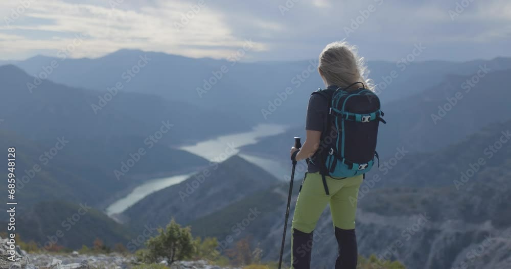 Active girl with hiking poles and backpack enjoying a view of river lake from high in the mountains. Standing on a cloudy and windy weather. Active girl with hiking poles and backpack enjoying a view of river lake from high in the mountains. Standing on a cloudy and windy weather.