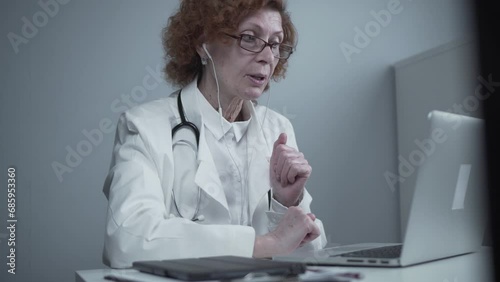 Female senior doctor therapist in white coat and headset talking video conferencing on laptop in office using online video calling application. Remote medical care for patients, online consultation. photo