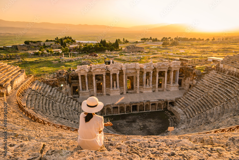 Hierapolis ancient city Pamukkale Turkey, a young woman with a hat watching the sunset by the ruins Unesco Heritage. Asian women watching the sunset at the old Amphitheater in Turkey during holiday - obrazy, fototapety, plakaty 