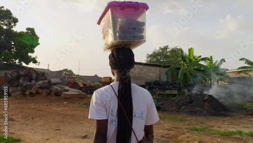Young woman seen from the back revealing her long beautiful hair, a plastic box on her head, walking through a sawmill compound, must be peddling drinks to the workers and the village, Kumasi, Ghana.