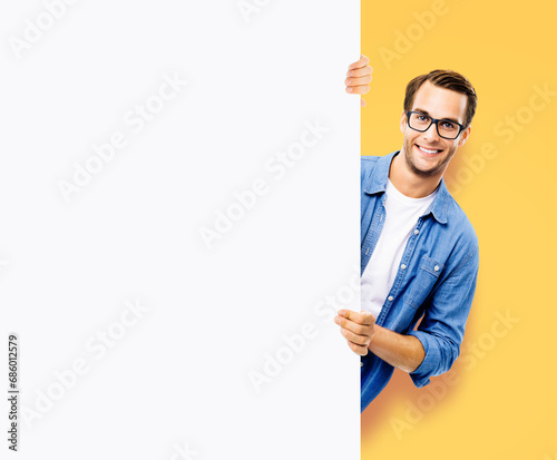 Portrait image - happy smiling casual man in eye glasses hold, peep out, stand behind mockup blank signboard, billboard, isolated orange yellow background. White empty ad sign board banner. Expert.