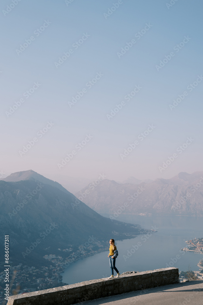 Young woman stands on a stone curb above the Bay of Kotor high in the mountains. Montenegro