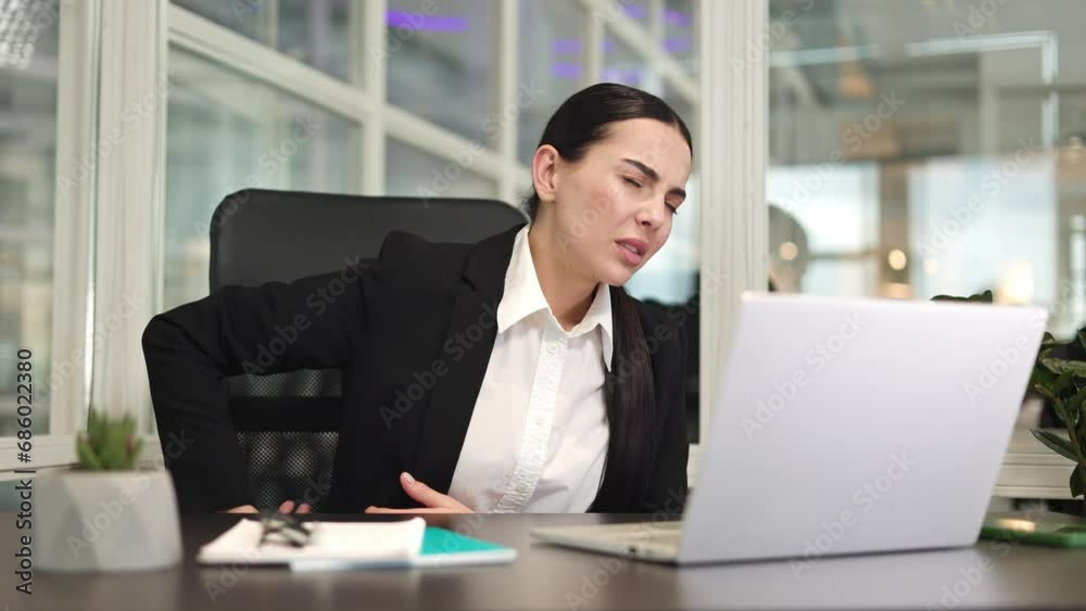 Sad business woman sitting at table and feeling side pain while working with digital computer in office. Caucasian adult employee putting hands below ribs and having hurt from working on device. Sad business woman sitting at table and feeling side pain while working with digital computer in office. Caucasian adult employee putting hands below ribs and having hurt from working on device.