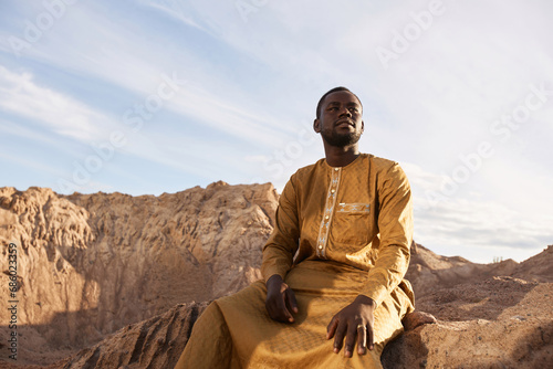 Canvas Print Dramatic portrait of Black young man wearing long kaftan sitting sand dune and l
