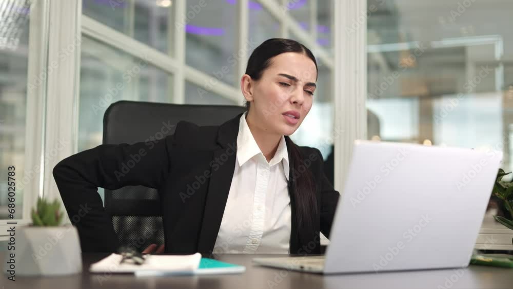 Concentrated female office worker sitting at desk and typing on wireless laptop keyboard while feeling back pain. Caucasian unwell lady trying stretching back at workplace in spacious room. Concentrated female office worker sitting at desk and typing on wireless laptop keyboard while feeling back pain. Caucasian unwell lady trying stretching back at workplace in spacious room.