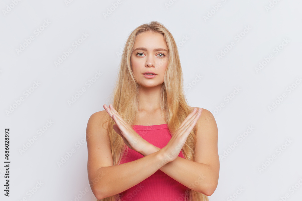 Serious blonde woman in pink top stands looking straight at camera with her arms crossed at chest in prohibitory sign, warning concept, copy space Serious blonde woman in pink top stands looking straight at camera with her arms crossed at chest in prohibitory sign, warning concept, copy space