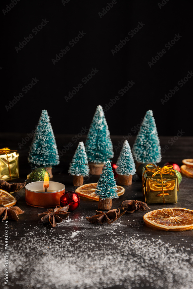 Top view of Christmas decoration with trees, candle, snow and orange on wooden table, black background, vertical, with copy space
