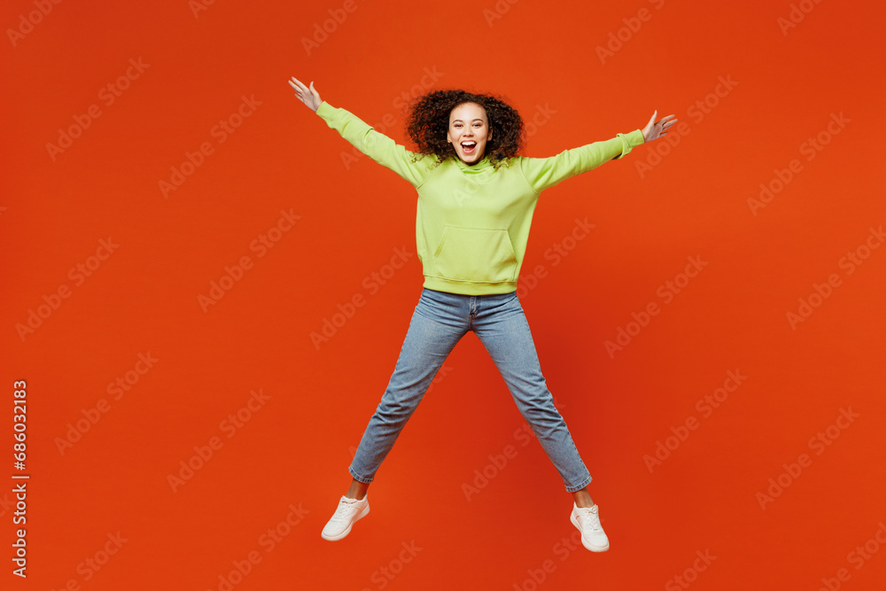 Full body happy young woman of African American ethnicity she wear green hoody casual clothes jump high with outstretched hands look camera isolated on plain red orange background. Lifestyle concept.