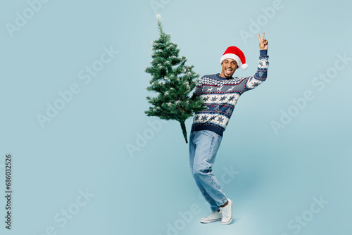 Full body side view fun young man wearing knitted sweater Santa hat posing hold in hand carry Christmas show v-sign isolated on plain blue background. Happy New Year 2024 celebration holiday concept.