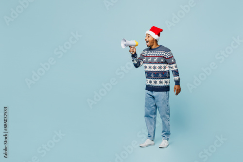 Full body young man wears sweater Santa hat posing scream in megaphone announces discounts sale Hurry up isolated on plain blue background. Happy New Year 2024 celebration Christmas holiday concept.