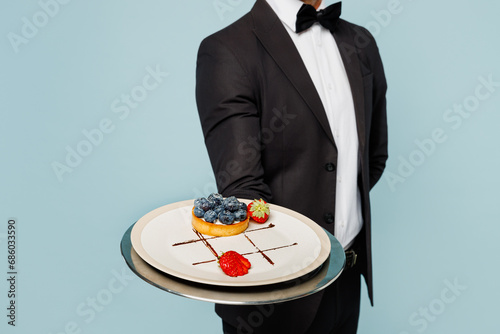 Cropped adult barista male waiter butler man wear shirt black suit bow tie elegant uniform hold show plate with cake order work at cafe isolated on plain blue background. Restaurant employee concept. photo