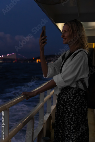 Young beautiful female tourist takes a video on a mobile phone while sailing on a ferry in the evening, selective focus. Concept of vacation, freedom, travel. Vertical photo