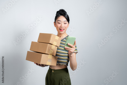 Young excited beautiful asian woman holding smartphone parcel cardboard standing on isolated white background. © makistock
