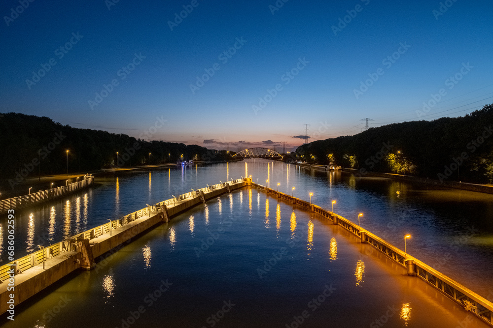 As twilight descends, this image showcases a river walkway illuminated by street lights, creating a path of light across the water. The walkway divides the river symmetrically, leading the eye towards